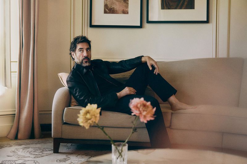Javier Bardem wearing a black leather jacket and trousers, reclining barefoot on a beige sofa in an elegant interior.
