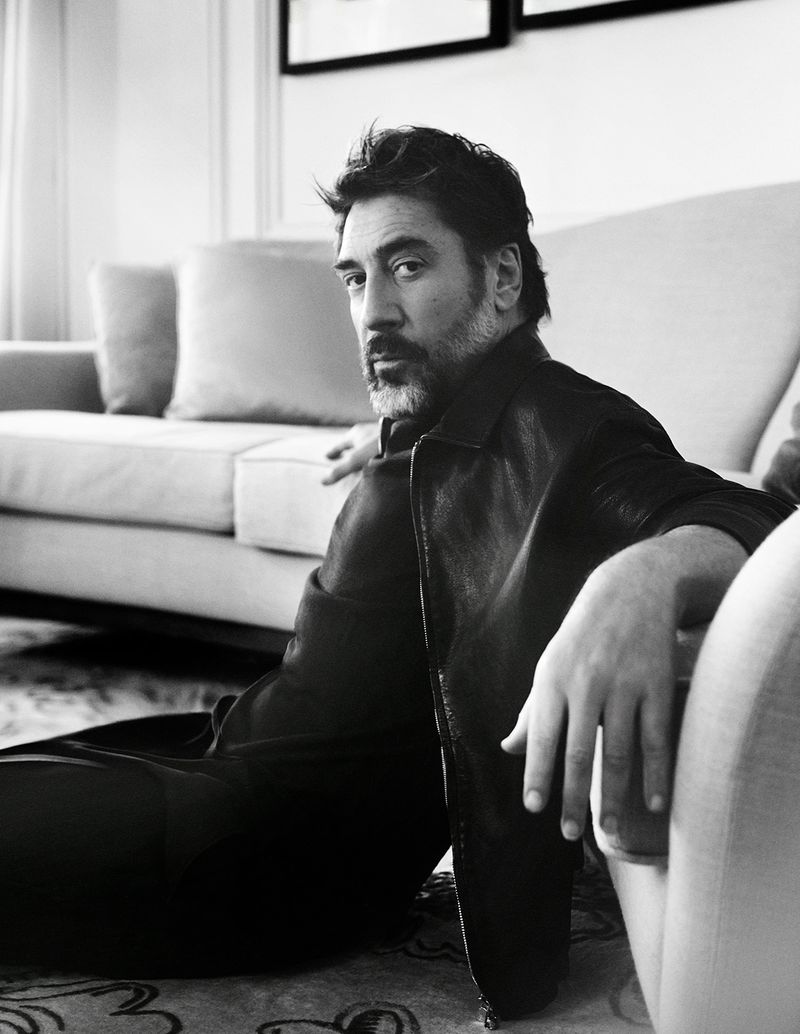 Actor Javier Bardem seated on the floor leaning against a sofa in a dark jacket for a black and white editorial photoshoot.