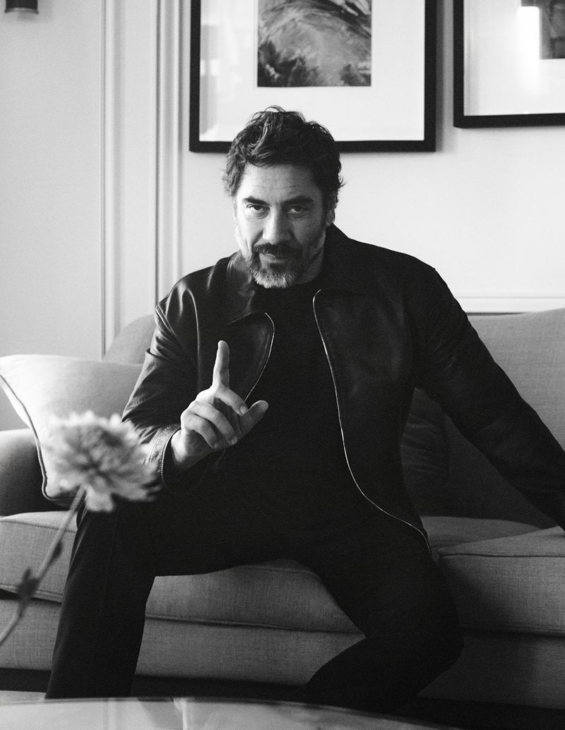 Actor Javier Bardem wearing a dark jacket seated on a sofa indoors in a black and white portrait.
