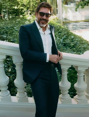 Actor Javier Bardem smiles wearing a dark suit, white unbuttoned shirt, and sunglasses while leaning against a white stone balustrade.