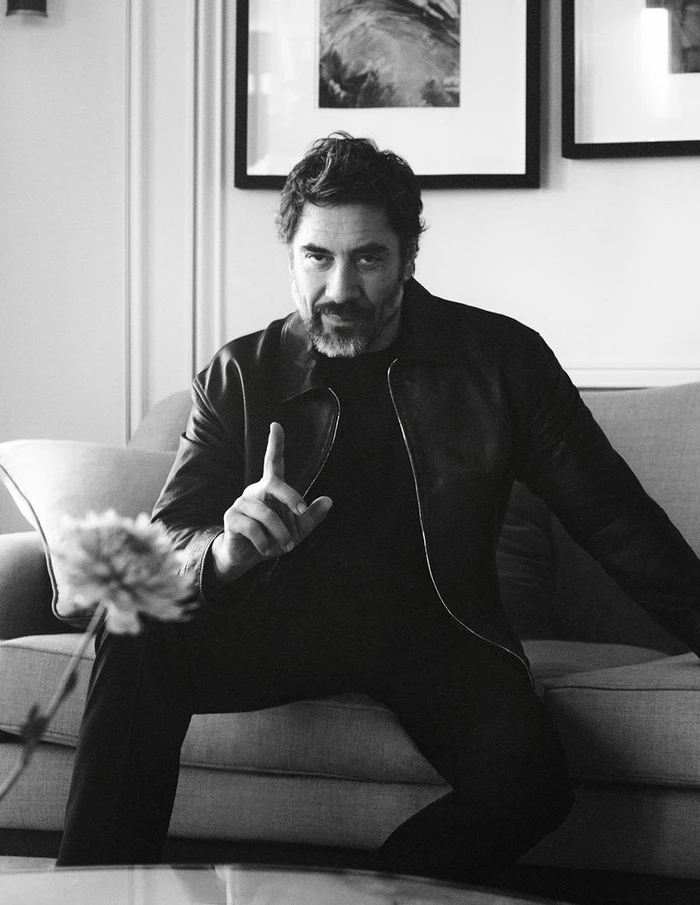 Actor Javier Bardem wearing a dark jacket seated on a sofa indoors in a black and white portrait.