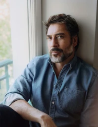 Actor Javier Bardem wearing a blue denim button-down shirt seated by a window frame in soft natural daylight.