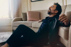 Actor Javier Bardem laughing while sitting on the floor leaning against a beige sofa in a sunlit room.