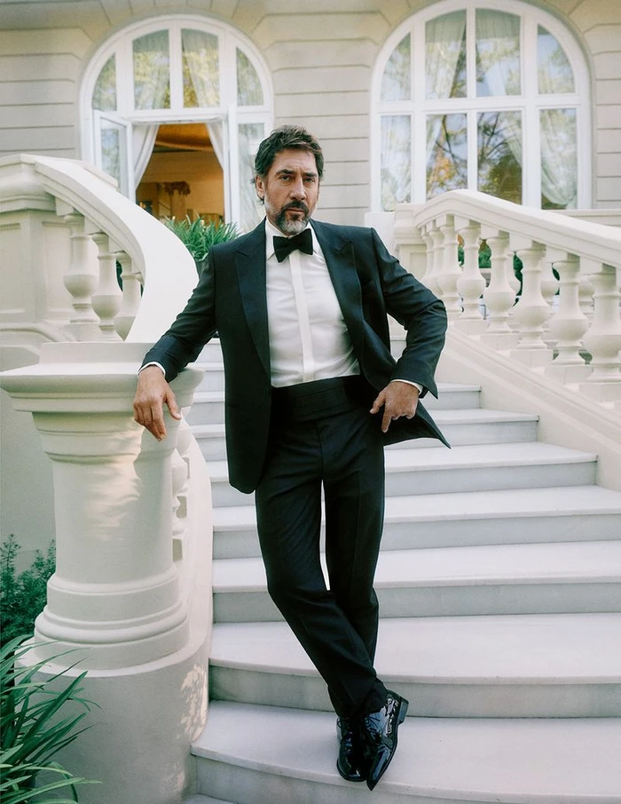 Actor Javier Bardem posing in a tailored black tuxedo with a black bow tie on an outdoor stone staircase.
