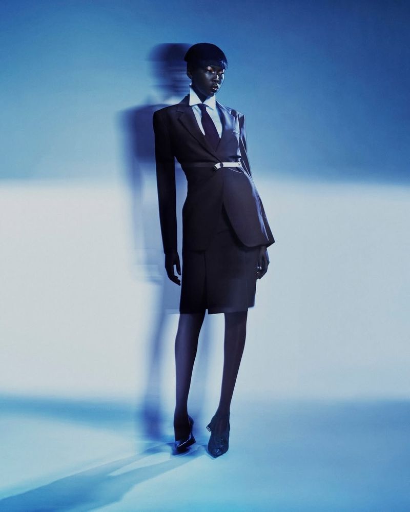 Model in a tailored black suit, white shirt, and tie standing against a cool blue studio background with motion blur shadows.