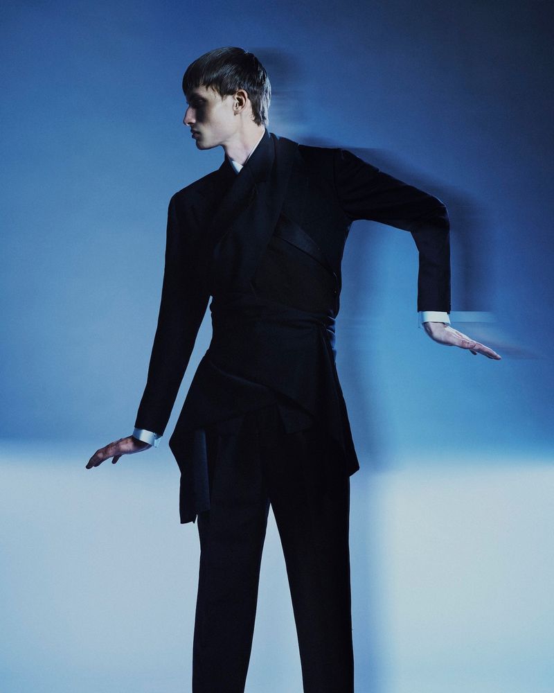 Male model in a structured black belted suit posing dynamically against a cool blue background with motion blur.