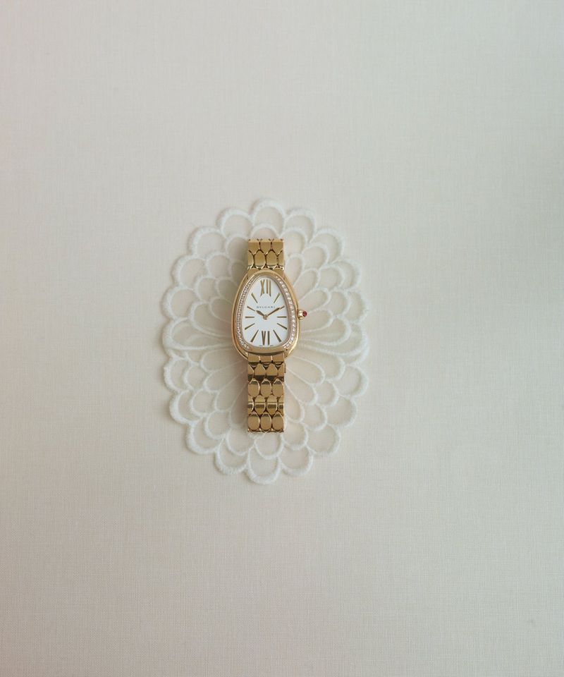 Gold Bvlgari luxury watch with a diamond bezel placed on a white ornamental lace cut-out against a clean off-white background.
