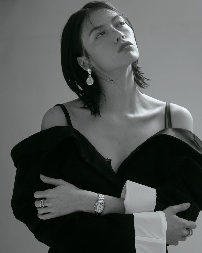 Lisa Han posing in an off-the-shoulder black coat with white cuffs, wearing a luxury watch and earrings against a seamless backdrop.