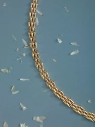 Gold geometric studded link necklace curving diagonally across a muted blue surface scattered with small white flower petals.