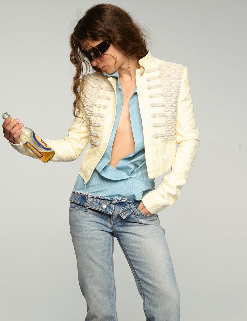 Model wearing an embellished cream military jacket, unbuttoned blue shirt, and low-rise denim jeans while holding a bottle.