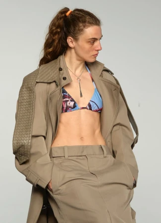 Model wearing an open oversized beige trench coat and printed blue bikini top against a grey studio backdrop.