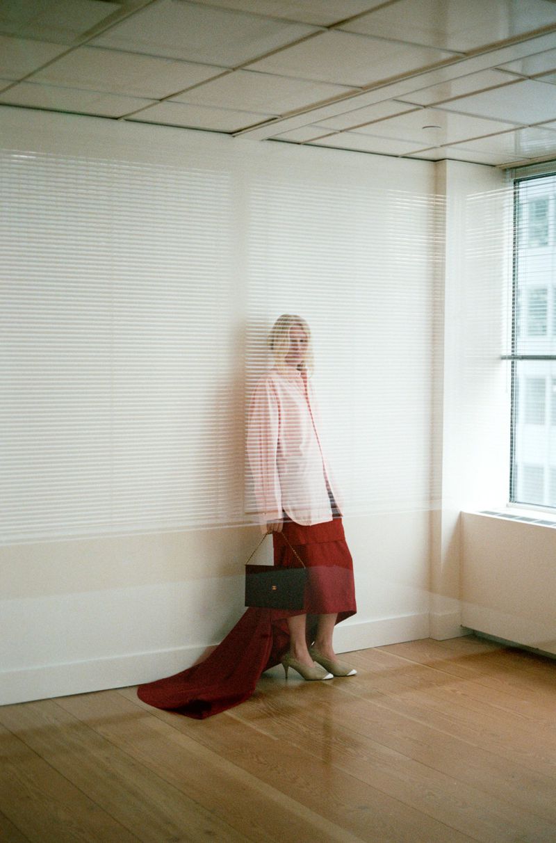 Model in a long collared shirt and trailing burgundy skirt holding a dark handbag in an empty office setting with window reflections.