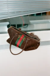 Brown suede designer handbag with green and red webbing stripe resting on a white windowsill beneath venetian blinds.