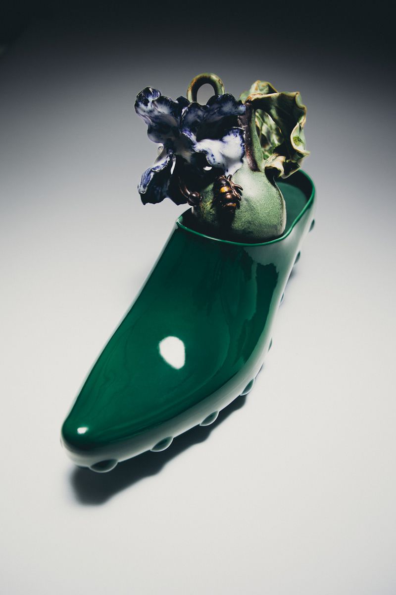 Glossy dark green slip-on clog shoe holding a sculpted ceramic floral arrangement against a neutral studio backdrop.