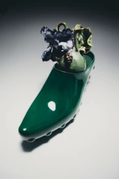Glossy dark green slip-on clog shoe holding a sculpted ceramic floral arrangement against a neutral studio backdrop.