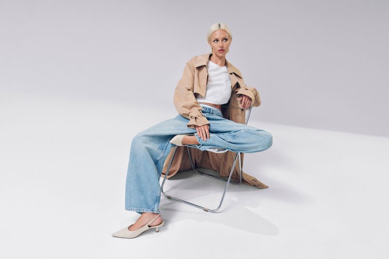 A fair-skinned woman with blonde hair in a trench coat and wide-leg jeans sits on a chair in a minimalist white photo studio.