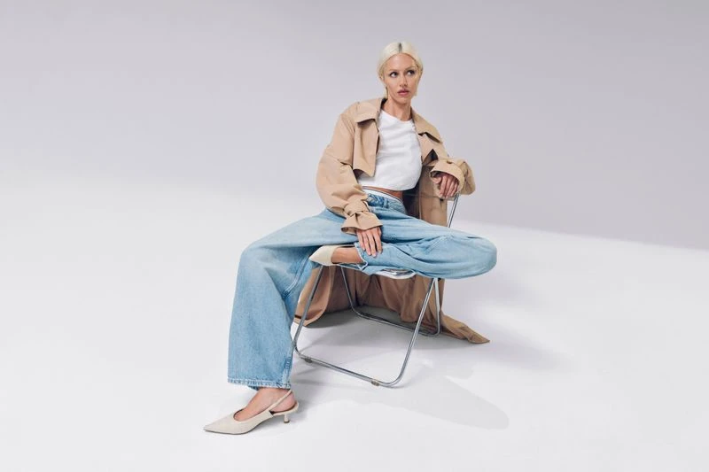 A fair-skinned woman with blonde hair in a trench coat and wide-leg jeans sits on a chair in a minimalist white photo studio.