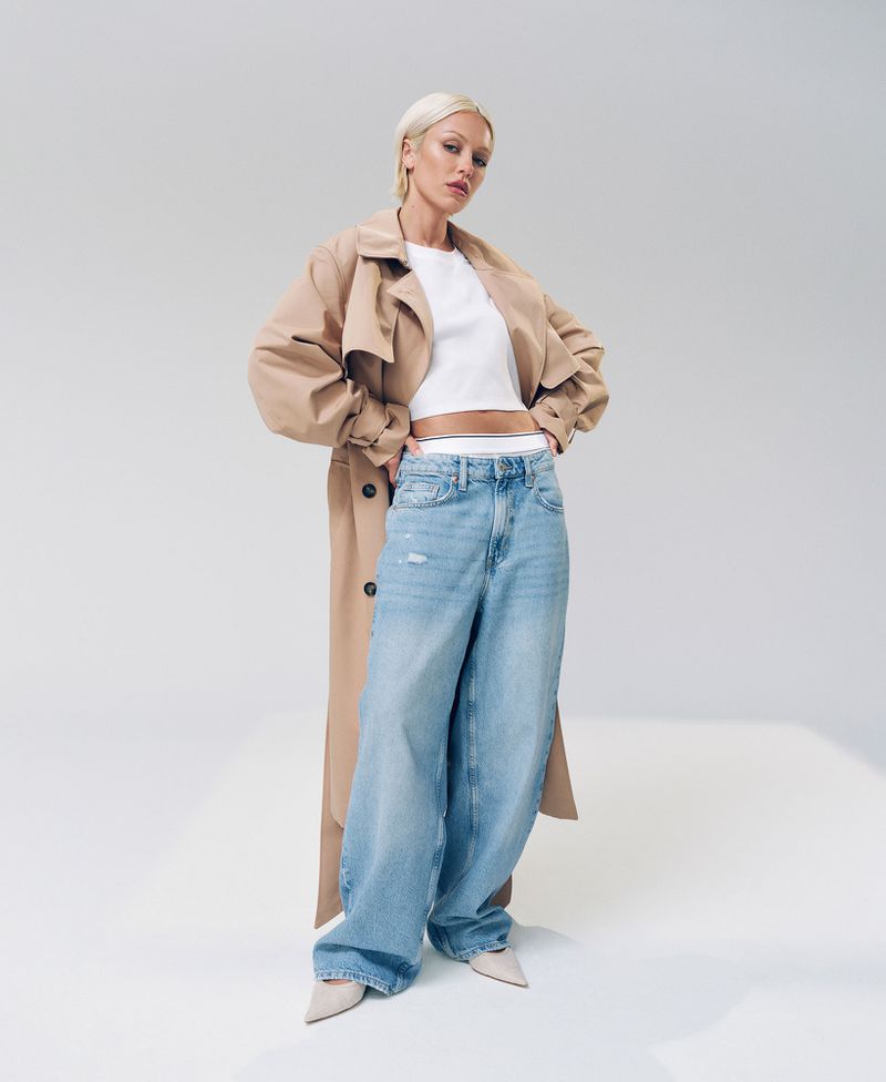A woman poses in light-wash wide-leg jeans, a white t-shirt, and a tan trench coat against a clean white backdrop in a fashion shot.