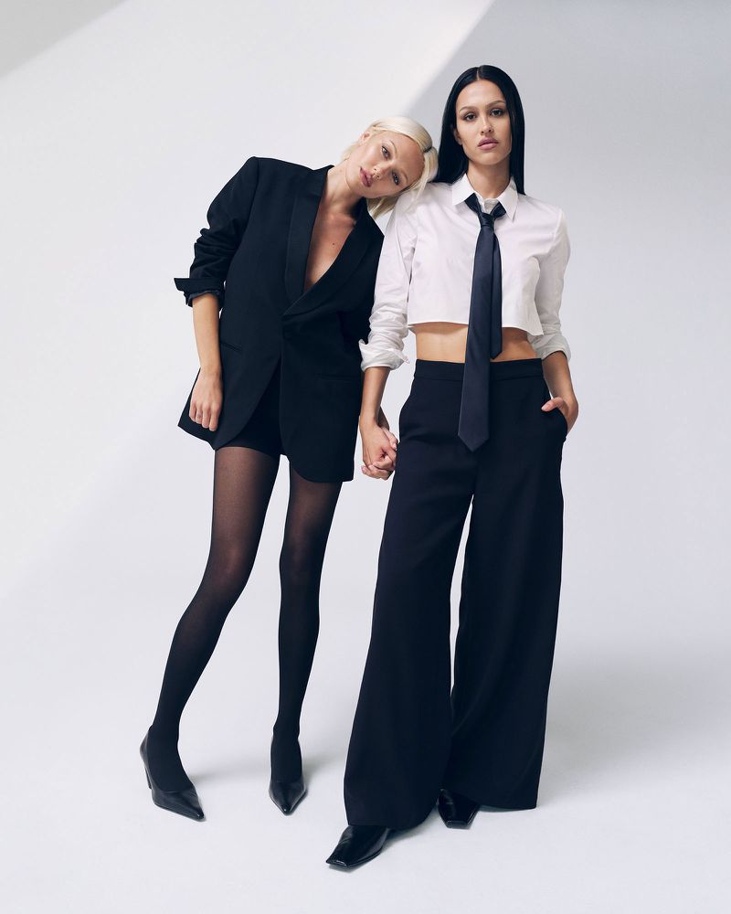 Two women pose in fashionable black and white outfits in a studio setting, one leaning her head on the other's shoulder.