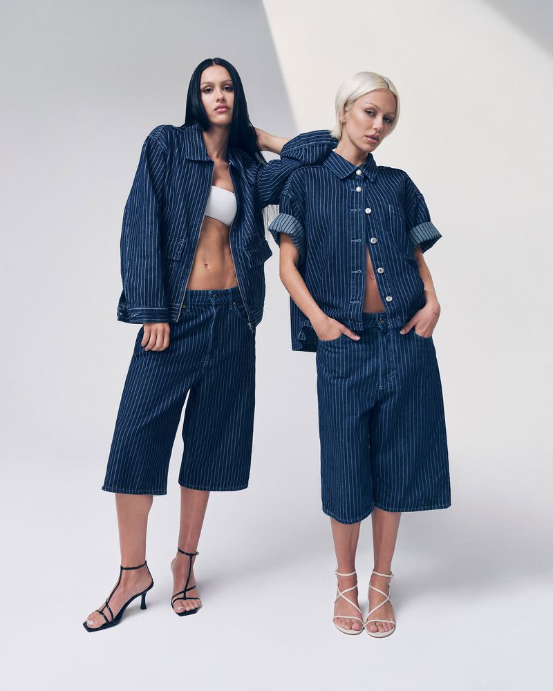 Two women pose in matching blue pinstripe denim outfits and strappy heels against a bright white background in a fashion studio shoot.
