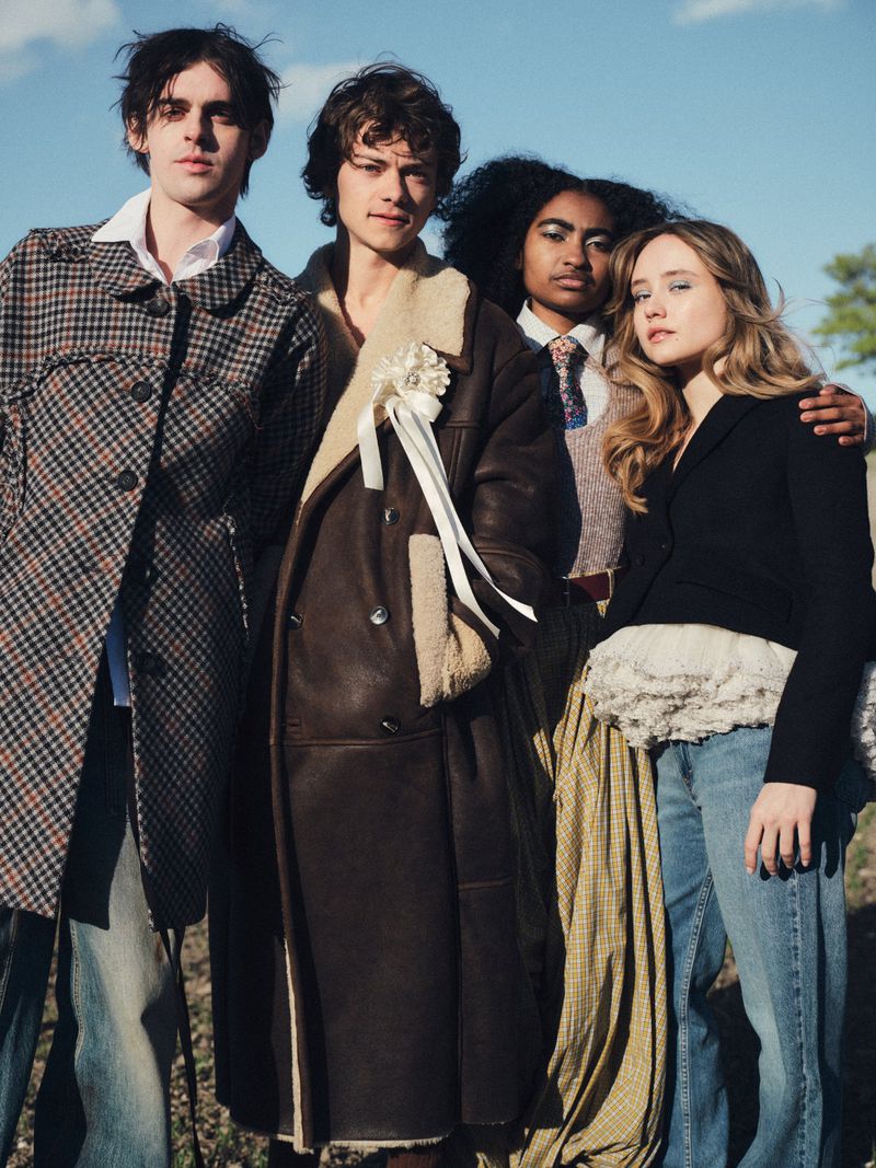Bella Ramsey and three models posing together outdoors against a blue sky wearing designer coats, jackets, and denim.