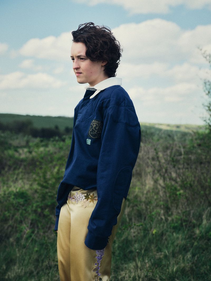 Actor Bella Ramsey stands outdoors in a grassy meadow wearing a navy collared shirt and embroidered gold trousers.
