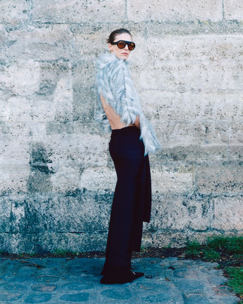 Model wearing dark sunglasses and a pale textured fringed wrap with black trousers against a stone wall.