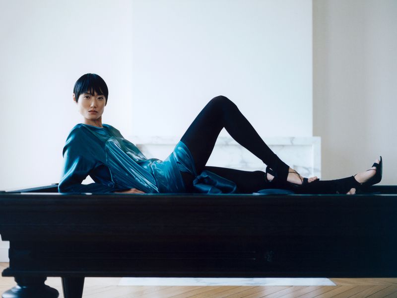 Model Ash Foo reclined on a dark wooden table wearing a shimmering teal top and black tights in front of a white fireplace.