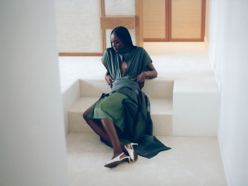 Model Ash Foo in a draped green dress and white slingback heels seated on minimalist carpeted steps indoors.