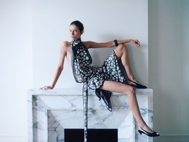 Model Martyna Budna posing in a patterned draped dress while seated atop a white marble fireplace.