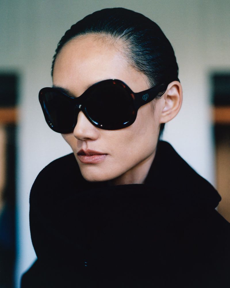 Model Ash Foo wearing oversized black sunglasses and a high-neck black coat against a soft-focus background.