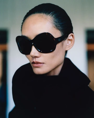 Model Ash Foo wearing oversized black sunglasses and a high-neck black coat against a soft-focus background.