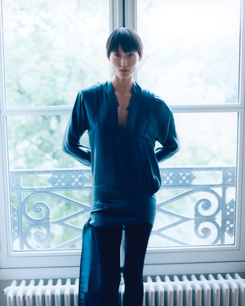 Model Ash Foo wearing a teal satin v-neck tunic and black leggings posed in front of a bright French window.