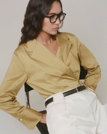 Model Summer Thompson seated wearing black glasses, an ochre wrap blouse, and white pants against a seamless studio background.