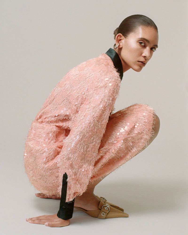Summer Thompson crouching in a textured blush pink dress and camel pointed buckle mules against a neutral studio backdrop.
