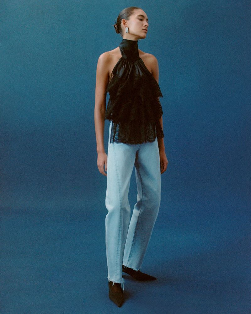 Model Summer Thompson wearing a high-neck black ruffled halter top and light-wash straight-leg jeans against a blue seamless backdrop.
