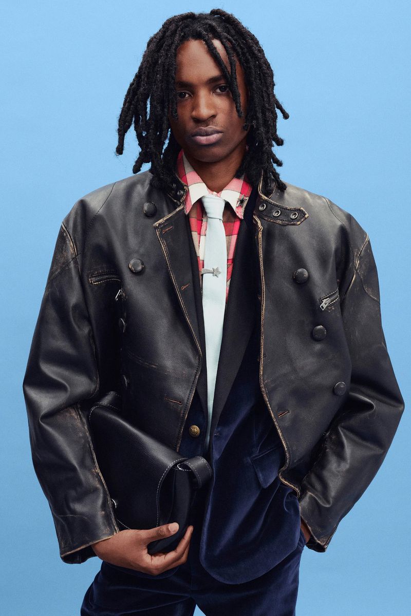 Black male model with dreadlocks wearing a distressed black leather jacket, plaid shirt, and tie against a light blue studio background.