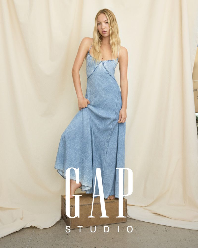 A blonde woman in a light blue denim dress stands barefoot on a wooden box in a bright studio setting with a cream backdrop.