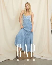 Model Lila Moss wearing a blue denim slip dress standing on a wooden crate against a cream fabric backdrop for Gap.
