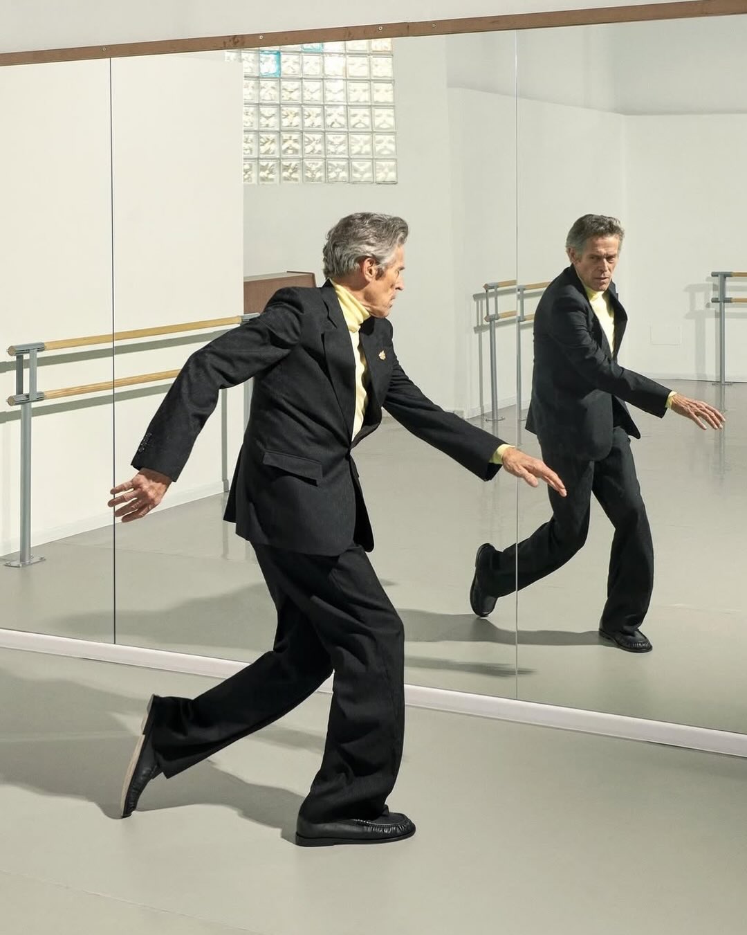 Actor Willem Dafoe wearing a black suit and yellow turtleneck while posing mid-movement in front of a dance studio mirror.