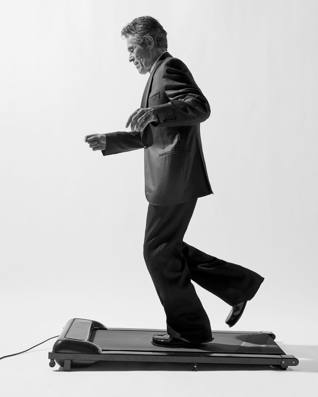 Willem Dafoe walking on a treadmill wearing a dark suit against a white studio background