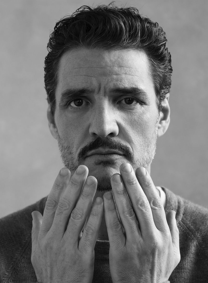 Black and white portrait of Pedro Pascal holding his hands up in front of his face against a smooth studio backdrop.