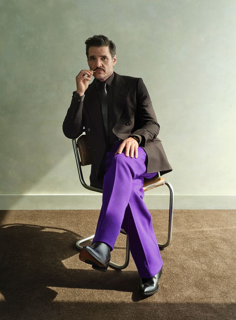 Pedro Pascal in a black suit jacket with purple pants, sitting on a chair in a studio with neutral wall and carpet background.