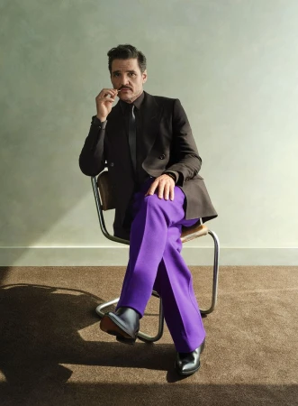 Pedro Pascal in a black suit jacket with purple pants, sitting on a chair in a studio with neutral wall and carpet background.