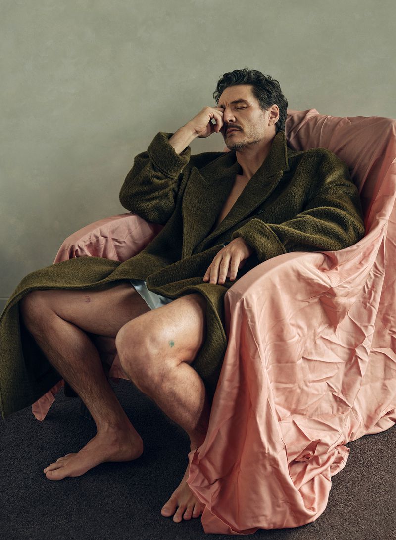 Adult male in olive robe sits in pink chair, indoors. He is barefoot, with eyes closed and hand to his face, resting or thinking.