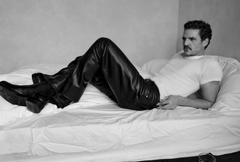 Portrait of a man lying on a white bed in a white t-shirt and black leather pants, posing casually with a neutral facial expression.