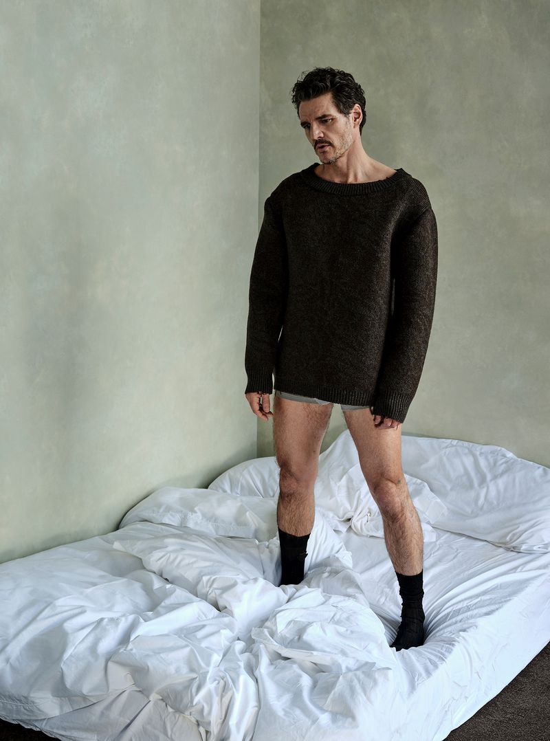 Man wearing sweater and socks stands on bed with rumpled white linens. He has dark hair, and a somber expression, against a neutral wall.