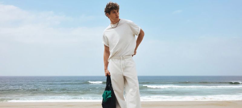 Model Valentin Humbroich wearing a white t-shirt and matching relaxed pants while standing on a beach holding a bag.