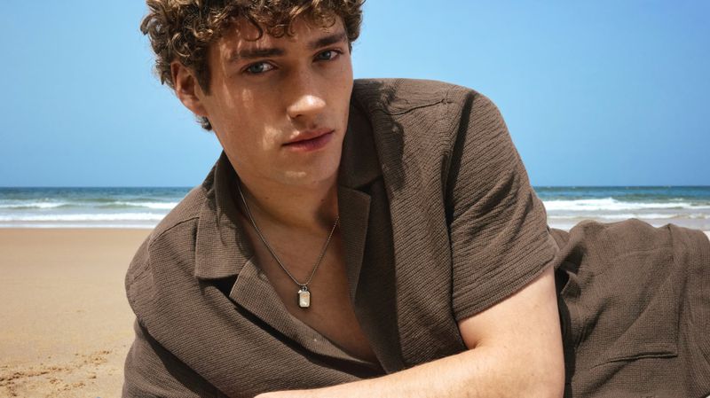Valentin Humbroich reclining on a sandy beach wearing an open-collar brown textured shirt and silver pendant necklace.