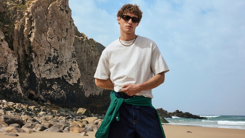 Model Valentin Humbroich in a white t-shirt and dark sunglasses standing on a rocky beach near cliffs.